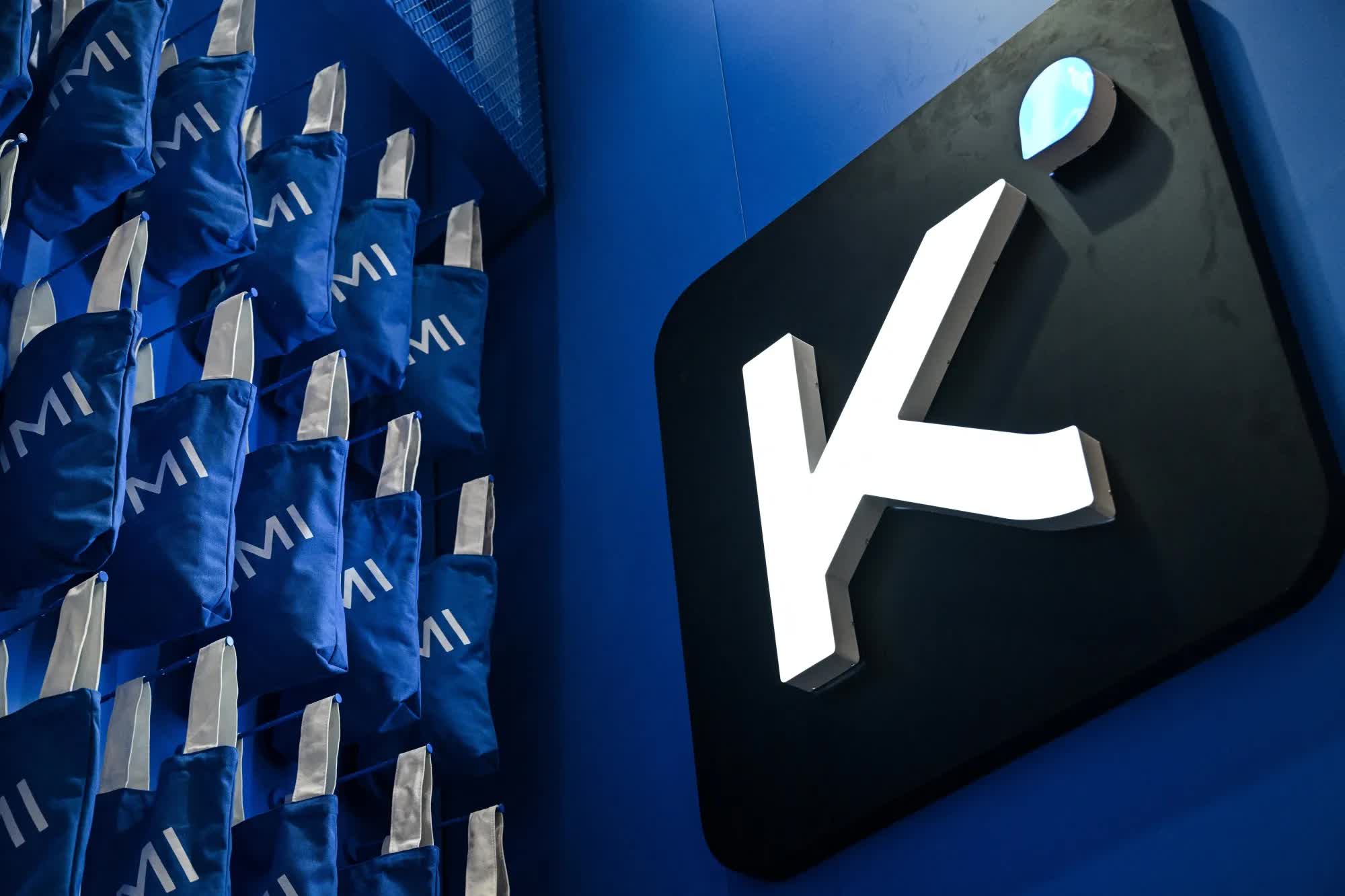 Illuminated Kimi logo mounted on a blue wall beside rows of branded tote bags at a Moonshot AI event